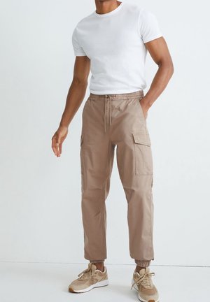 Hombre con camiseta blanca de manga corta, pantalones cargo beige con puños elásticos y zapatillas marrones, de pie frente a un fondo blanco.