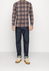 Mężczyzna w beżowej i czarnej kraciastej koszuli z guzikami, ciemnoniebieskich dżinsach z podwiniętymi nogawkami oraz beżowych zamszowych butach na grubej podeszwie.