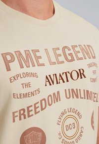 Camiseta de algod�ón beige con texto impreso destacado en marrón, que incluye "PME LEGEND", "AVIATOR" y varios logotipos y frases.