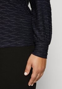 Marinblus med strukturerat vågmönster och metalltrådar, med långa ärmar med gummiknappar, ihop med ett svart plagg.