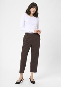 Weißes Langarmoberteil, braune hochtaillierte Hose mit Falten und schwarze Slingback-Absätze. Glatter, enganliegender Stoff, gerades Bein-Design.