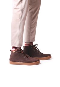 Zapatos de ante marrón de caña alta con suelas de goma marrón, que presentan un diseño de cordones y calcetines a rayas en rojo y beige.