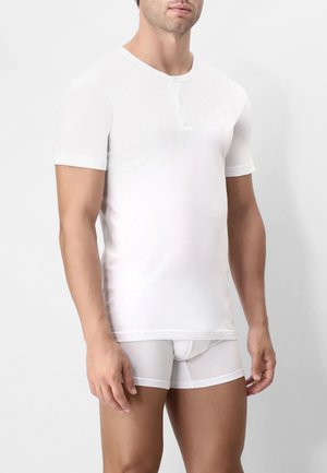 Uomo che indossa una semplice maglietta henley bianca a maniche corte e slip bianchi, in piedi contro uno sfondo chiaro.