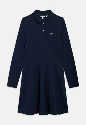 Temno moder polo kroj z dolgimi rokavi, izdelan iz mehke tkanine. Ima ovratnik in zelen Lacoste logotip na prsih. Krilo je razširjeno.