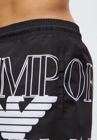 Čierne šortky z hladkej látky s veľkým bielym grafickým dizajnom s textom a logom s krídlami, elastickým pásom a bočným vreckom.