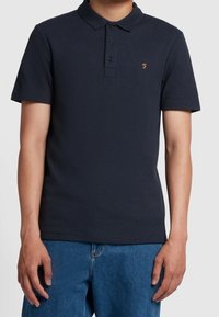 Marineblaues Poloshirt mit kurzen Ärmeln, gerippter Textur, Drei-Knopf-Leiste und einem kleinen orangefarbenen Logo auf der linken Brust, kombiniert mit Jeansshorts.