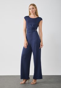 Navyblå jumpsuit med axelärmar, figursydd bodice och vida ben. Har en vriden framtill detalj och guldtonad accent vid midjan.