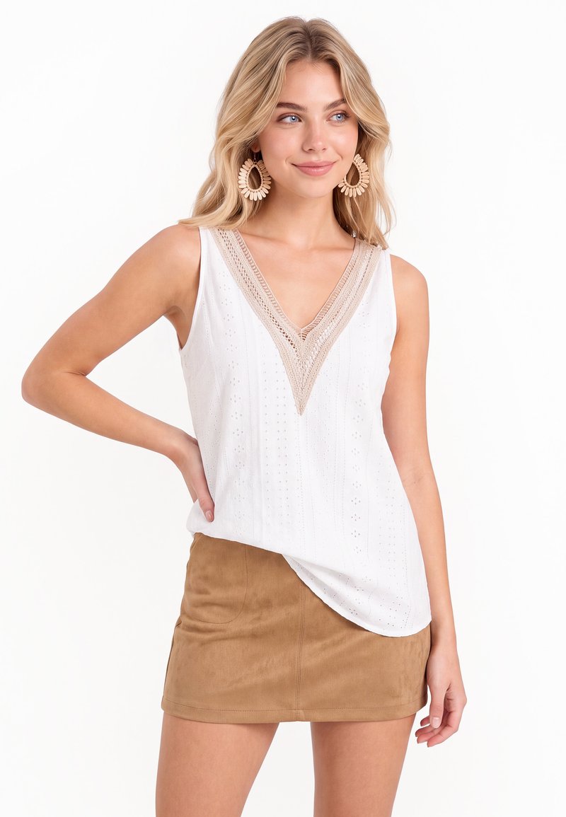 Femme blonde portant un haut blanc sans manches avec un col en V en dentelle beige et une mini-jupe en daim marron clair, souriante et regardant vers la droite.