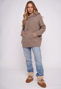 Brązowy dziergany sweter z kapturem, sznurkiem i przednimi kieszeniami, w połączeniu z jasnoniebieskimi dżinsami i beżowymi butami wyściełanymi futerkiem.