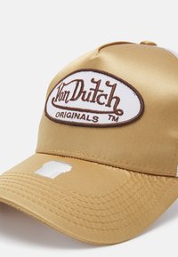 Zlatý saténový baseballový klobouk s bílou oválnou nášivkou s nápisem "Von Dutch Originals" v hnědé barvě. Obsahuje nastavovací pásek a prošívané švy.