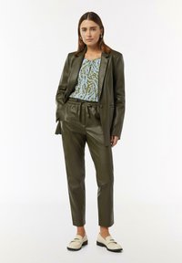 Olivgrüne Lederblazer und -hose mit einem Kordelzugbund, kombiniert mit einem blau- und grün gemusterten Oberteil und beigen Slippern.