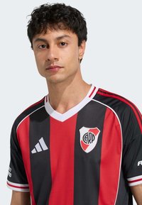 Camiseta de fútbol a rayas negras y rojas con cuello en V; presenta un logo en el pecho y tres acentos a rayas en las mangas.