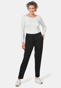 Zwarte broek met plooien en een comfortabele tailleband, gedragen met een witte top met lange mouwen. Model draagt witte sneakers.