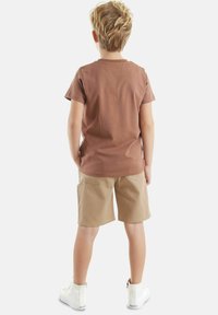 Een bruin katoen t-shirt en beige shorts met achterzakken, gecombineerd met witte enkellaarsjes, gezien vanaf de achterkant tegen een effen achtergrond.