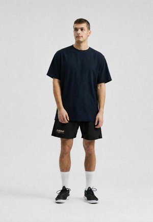Mladý muž stojaci, oblečený v čiernom oversized tričku, čiernych šortkách s logom, bielych členkových ponožkách a čiernych športových topánkach na svetlosivom pozadí.