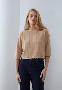 Beige Rippenstrickoberteil mit kurzen Ärmeln und einem rohen Saum, kombiniert mit dunkelblauen Jeans im klassischen Fünf-Taschen-Stil.