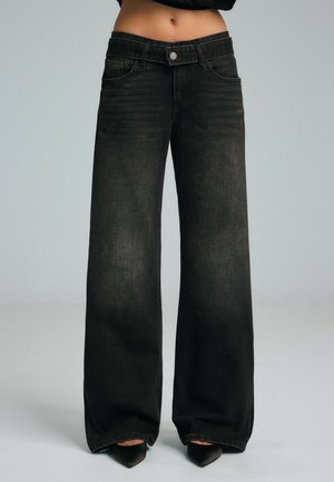 Femme portant un jean en denim noir taille haute à jambes larges et des chaussures noires à bouts pointus, debout devant un fond gris uni.