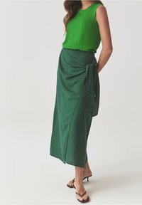 Donna che indossa un top verde senza maniche e una gonna a portafoglio verde scuro con dettaglio a fiocco, abbinati a sandali con tacco marroni.