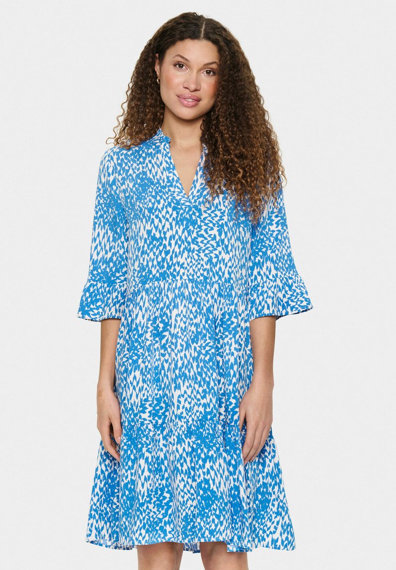Robe imprimée bleue et blanche avec décolleté en V, manches trois-quarts en cloche, et ourlet évasé, fabriquée à partir d'un tissu léger avec une texture fluide.