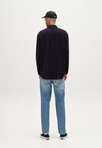 Camisa de pana morada, de manga larga, con pliegue en la parte posterior, puños abotonados, combinada con jeans de mezclilla azul claro y zapatillas negras. Fondo minimalista.