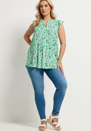 Femme portant un chemisier blanc sans manches à imprimé floral vert, un jean skinny bleu, des sandales blanches, ainsi que des boucles d'oreilles et des bracelets dorés.