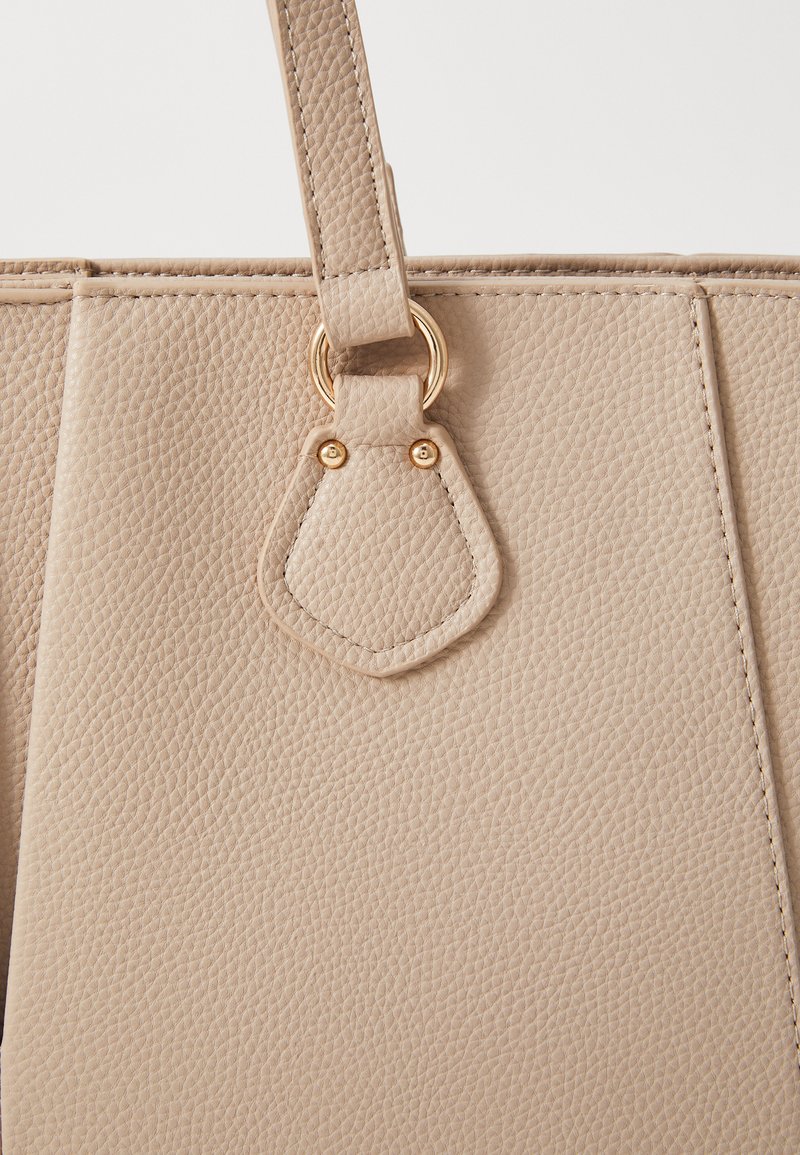 Bolso de mano de cuero texturizado beige con un amplio compartimento principal, que presenta un acento de anillo en tono dorado y un enganche de correa a juego.