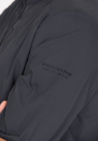 Tummanharmaa takki, jossa on kevyt, tekstuuri kuitu ja sileä pinta. "KESTÄVYYS TIEN SUURI HERO" painettuna hihaan.