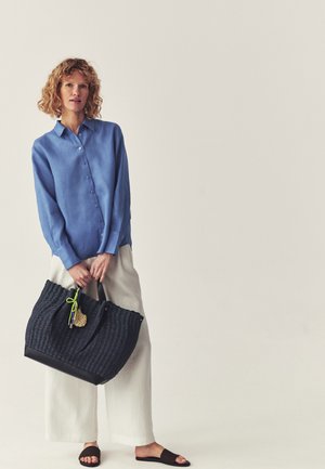 Frau mit lockigem Haar, die ein blaues Hemd mit Knöpfen, weiße weit geschnittene Hosen, schwarze Slide-Sandalen trägt und eine große dunkel geflochtene Tragetasche hält.