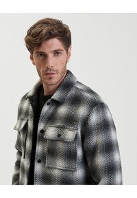 Giubbotto di lana a scacchi bianco e nero con colletto, due tasche frontali e dettagli dei bottoni in vista. Tessuto texture con motivo a spina di pesce.