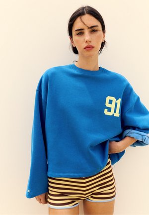 Junge Frau mit dunklem Haar trägt einen blauen übergroßen Pullover mit der Nummer 91 und gestreifte braune und gelbe Shorts vor einfarbigem Hintergrund.
