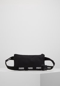Svart tygpåse med dragkedja, med ett tjockt bälte med "PUMA"-logotyp i vitt. Slät textur, kompakt rektangulär form.
