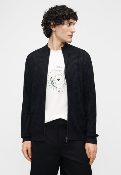 Άνδρας με σγουρά μαλλιά που φοράει μαύρη πλεκτή ζακέτα με φερμουάρ πάνω από λευκό T-shirt με γραφικά και μαύρο παντελόνι, κοιτάζει προς τα δεξιά.