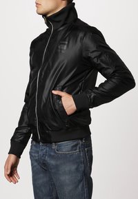 Svart bomberjacka av glänsande material, med hög krage, dragkedja, ribbade muddar och sidofickor, bärs över blå jeans.