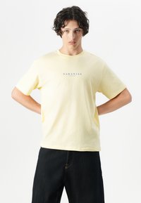 T-shirt in cotone giallo con maniche corte, caratterizzato da un logo blu "NARANJAS" al centro. La vestibilità è comoda, perfetta da abbinare a jeans scuri.