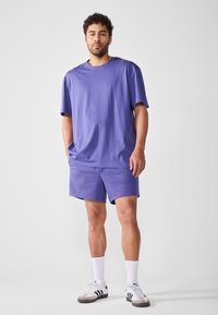 Camiseta de algodón púrpura y pantalones cortos a juego. Corte relajado con mangas cortas. Cuello acanalado notable. Calcetines blancos y zapatillas completan el look.