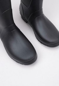 Bottes en caoutchouc noires avec une texture lisse, une pointe arrondie et une semelle striée. Présentent un design de hauteur intermédiaire pour protection en conditions humides.