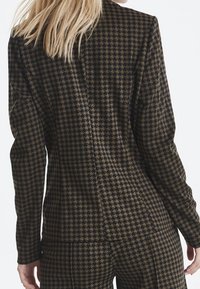 Kvinna i en kavaj och matchande byxor i brunt och svart hundtandmönster, sedd bakifrån med blont hår.