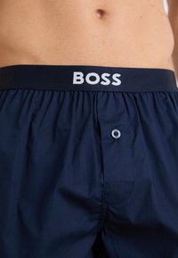 Mørkeblå svømmebukser med en hvit logo-belte, med knappelukking og rynket stoff. Glatt tekstur, uten synlige mønstre.