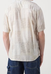 Kortærmet skjorte i lys beige stof med et paisley-mønster. Glat tekstur med en afslappet pasform og rund kant.