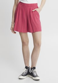 Rosa, leichte Shorts mit elastischem Bund und Seitentaschen, kombiniert mit schwarzen High-Top-Sneakern mit weißen Schnürsenkeln und Logo.