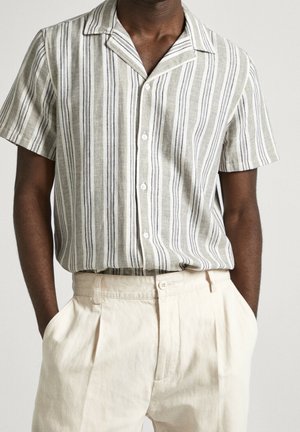 Mies, joka pukeutunut lyhythihaiseen, pystysuorilla harmailla ja valkoisilla raidoilla varustettuun napitettuun paitaan, joka on tungettu korkeavyötäröisiin, kermaan vivahtaviin laskostettuihin housuihin.