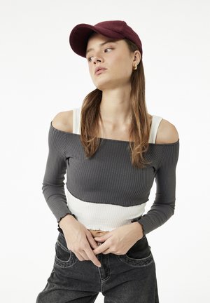 Jonge vrouw met lang haar die een bordeauxrode pet draagt, een grijze off-shoulder top over een witte tanktop en zwarte jeans, kijkt zijwaarts.