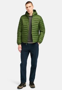 Groene geïsoleerde jas met een gestructureerde afwerking, ritsluiting en capuchon, gedragen over een grijze trui, gecombineerd met donkere broeken en stevige laarzen.