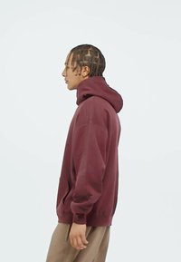 Ung person med flätat hår som bär en vinröd hoodie och beige byxor, stående i profil mot en enkel ljus bakgrund.