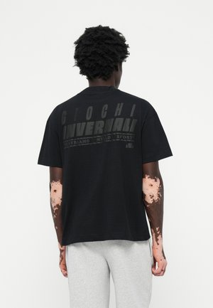 Sort kortærmet t-shirt med fed, struktureret tekst på ryggen. Stoffet ser blødt ud; afslappet pasform. Båret med lysegrå sweatpants.
