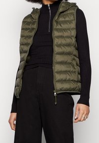 Olivgrüne gepolsterte Weste mit Kapuze, ausgestattet mit einem Reißverschluss vorne, horizontalem Steppdesign und einem gerippten schwarzen Rollkragenpullover darunter.