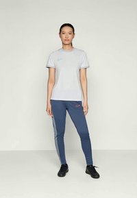 Šviesiai pilkas trumpomis rankovėmis sportinis marškinėliai su mėlynu Nike logotipu; derinami su tamsiai mėlynomis bėgimo kelnėmis, kurios turi raudoną Nike logotipą ir pilkas šonines juostas.
