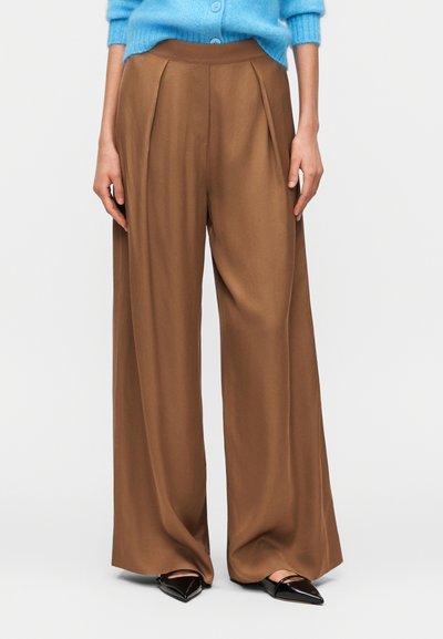 Personne portant un pantalon marron taille haute à jambes larges avec des plis, des ballerines noires à bout pointu, et un cardigan bleu court avec des boutons.