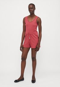 Combinaison en coton rouge avec col rond, sans manches, taille élastique à cordon de serrage et perles décoratives ; texture lisse et coupe décontractée.