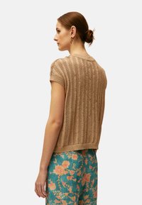 Mujer con el cabello recogido en un moño, lleva pendientes de aro dorados, una camiseta beige de punto de manga corta y pantalones de color azul verdoso con estampado floral, mirando hacia la izquierda.
